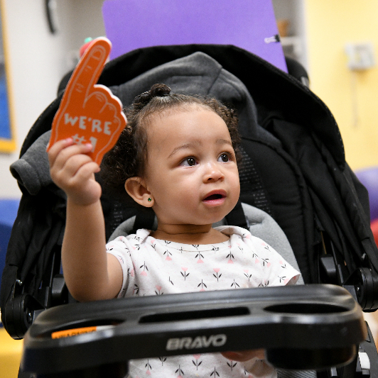 A child in a stroller holding an orange foam #1 finger. 