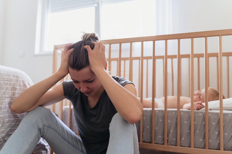 Woman sitting next to her child's crib with her head in her hands - feeling anxious and depressed in postpartum.
